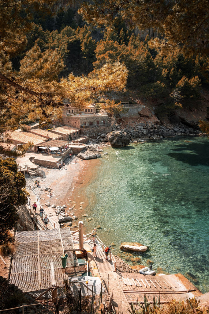 Cala Deià in Mallorca, a small pebble cove with clear turquoise water below stone boathouses and pine-covered cliffs