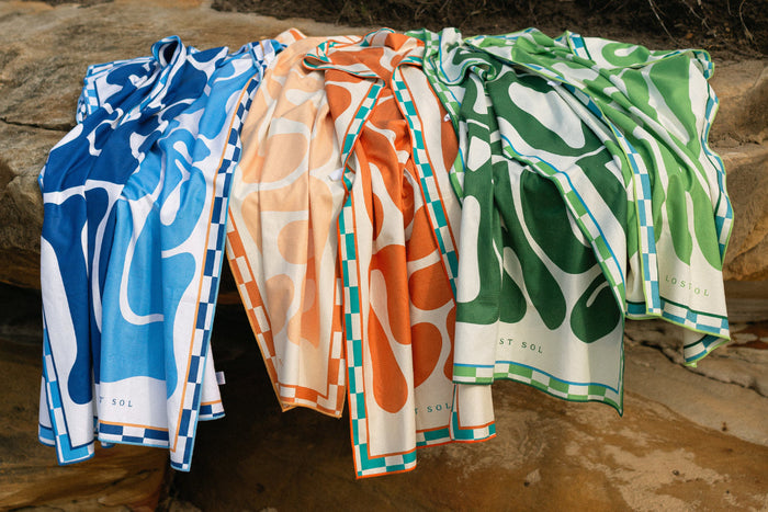 Lost Sol beach towels in blue, orange and green draped over rocks outdoors.