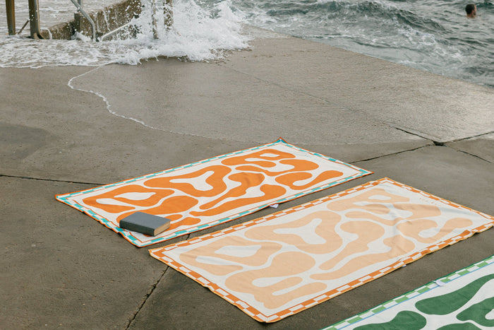 Two orange Mallorca beach towels, Bold and Pastel, laid out by the ocean on a concrete platform with waves crashing nearby.
